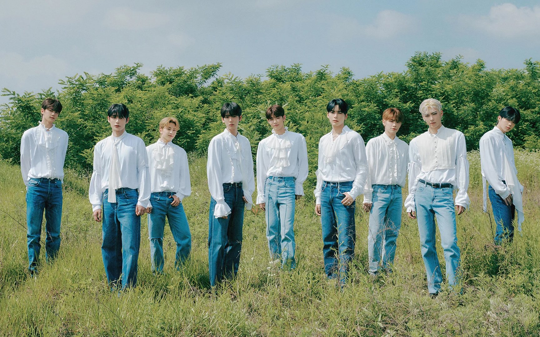 The nine members of ZEROBASEONE in white shirts and jeans, standing in front of a grass field and green woods — a Youth Trilogy era concept photo