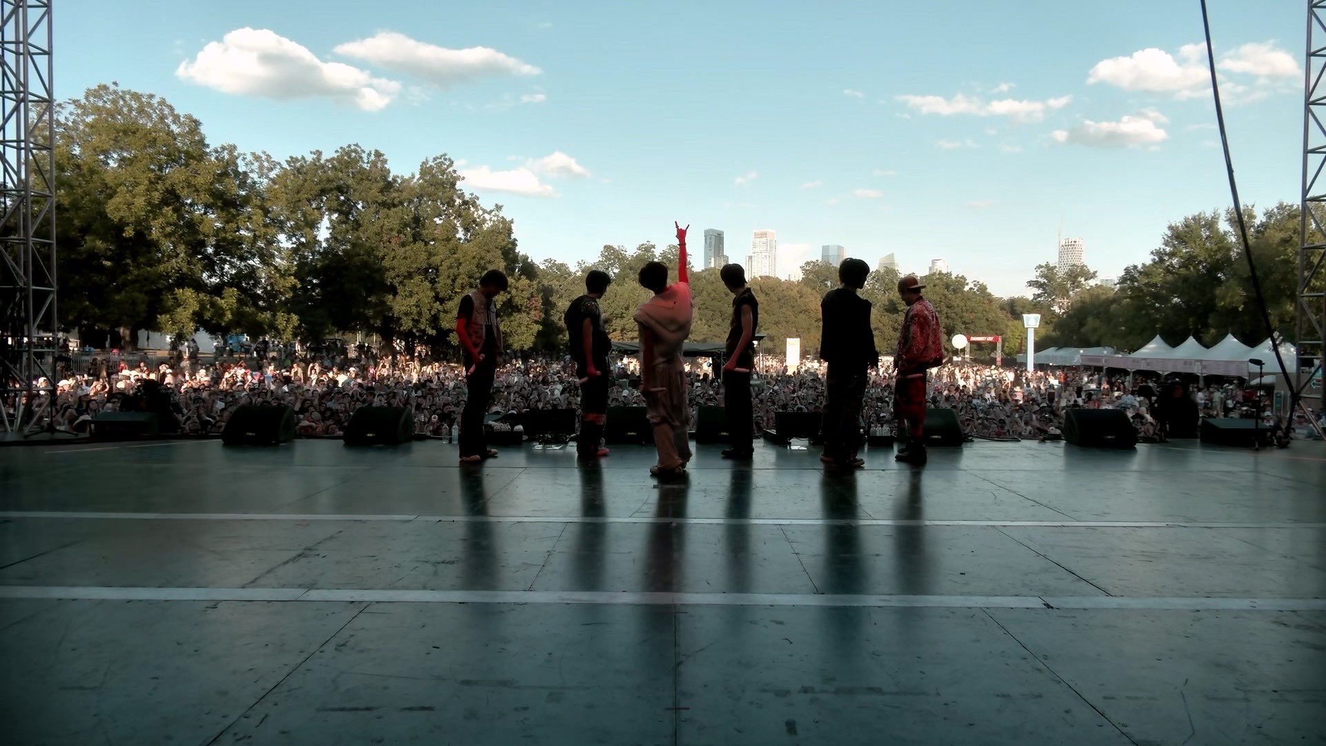 RIIZE on an outdoor festival stage before a global crowd with a city skyline behind them