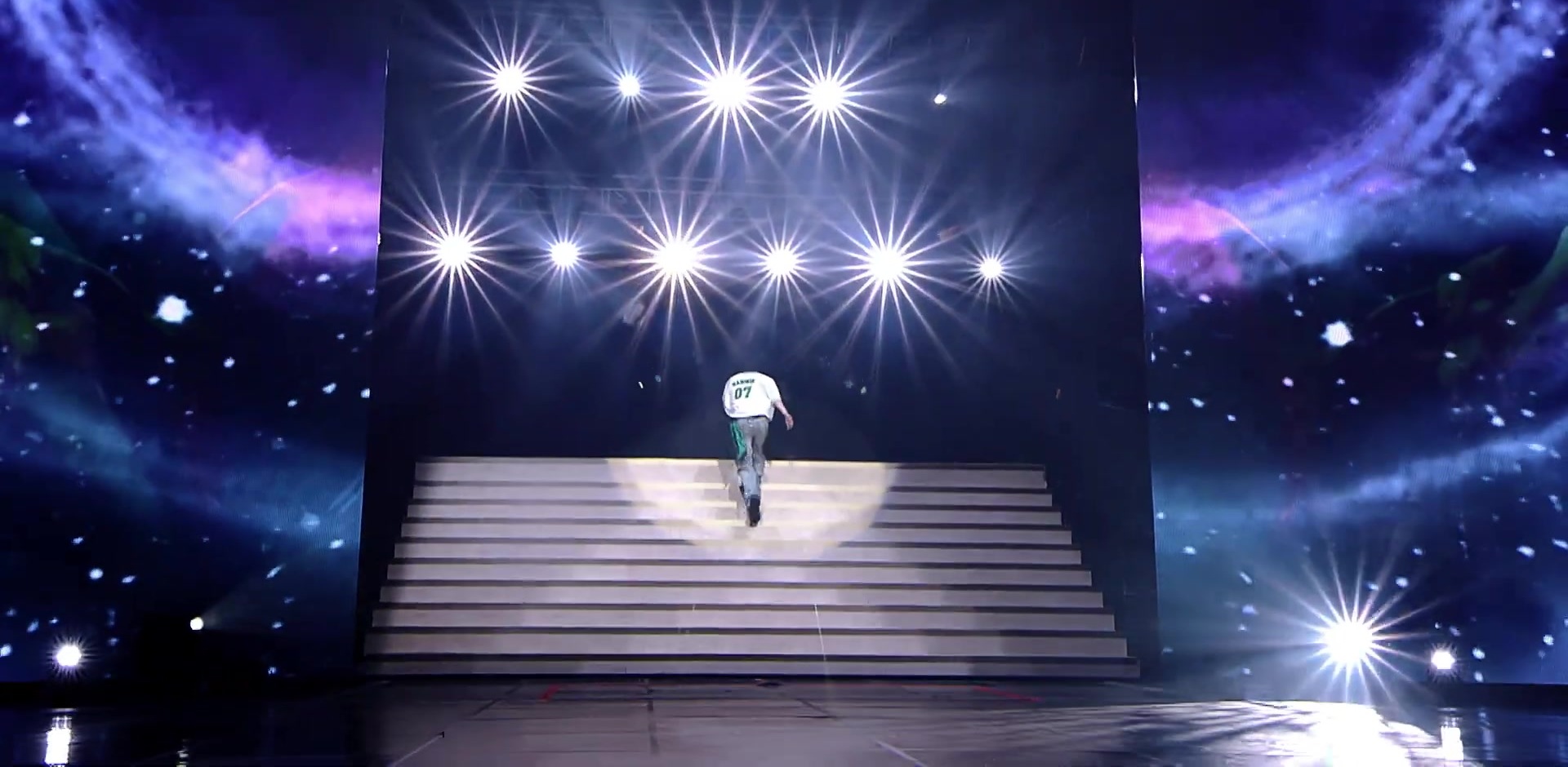 TAEMIN standing alone at the center of a staircase on his world tour stage as twelve lights and a space-themed screen face him