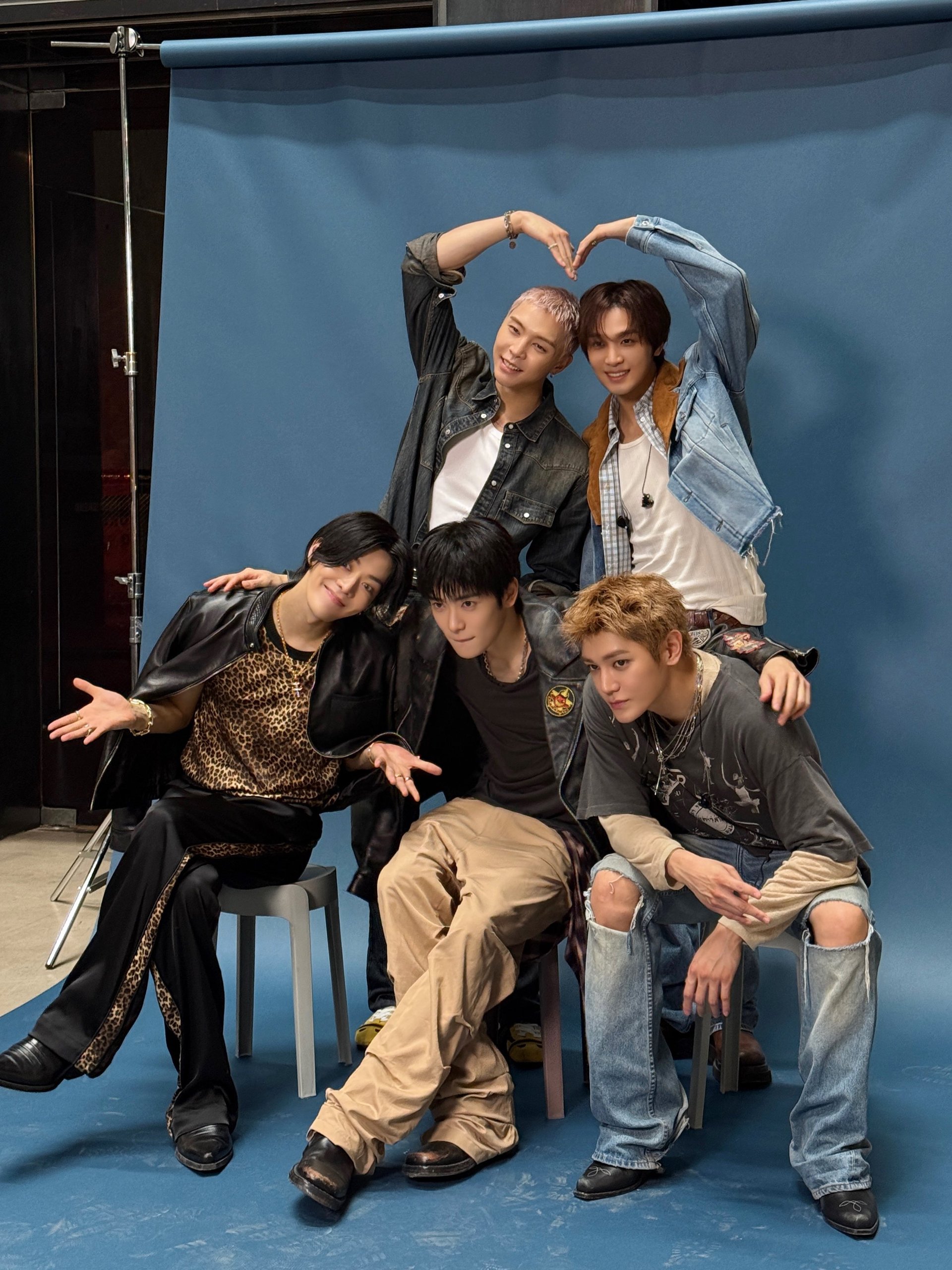 I cinque membri degli NCT 127 seduti insieme mentre formano cuori con le mani durante la promozione di BLINGY