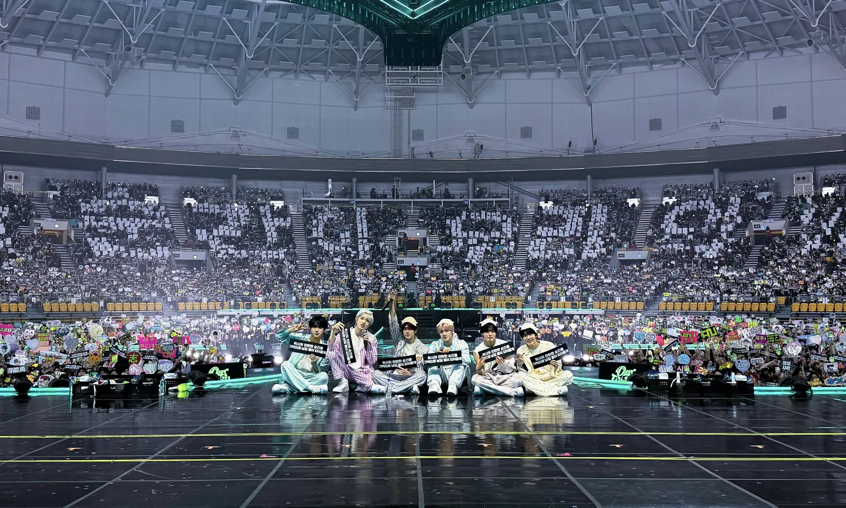 Шестеро участников NCT WISH сидят рядом на краю сцены KSPO DOME, а за ними трибуны, заполненные фанатами с плакатами поддержки, — финальный концерт INTO THE WISH в Сеуле